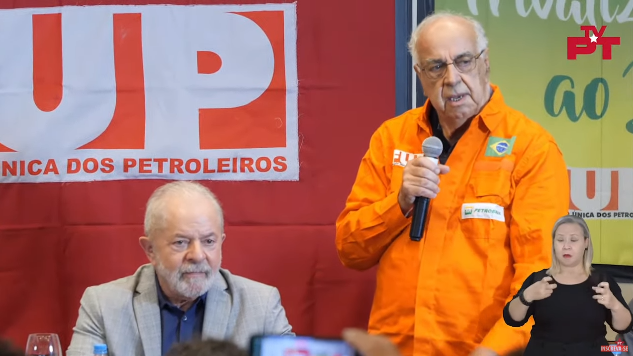 O ex-presidente Lula (PT) participando de debate com petroleiros. Ele está sentado, atrás dele há uma bandeira da FUP e ao seu lado um homem está em pé e fala ao microfone.