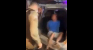 Homem sendo colocado no porta-malas da viatura por policiais.