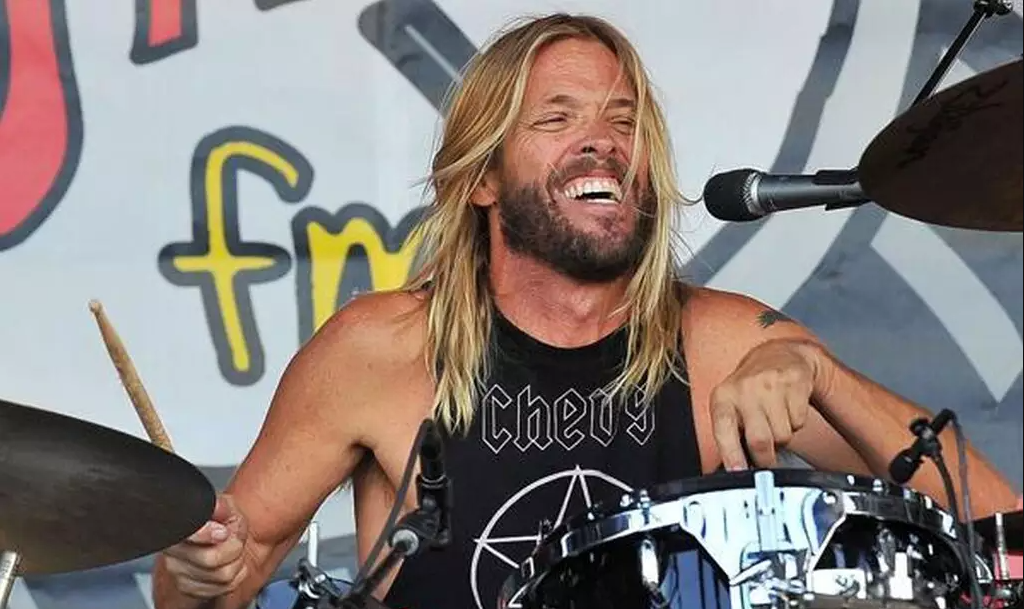 Imagem de Taylor Hawkins, de cabelo longo e louro, tocando bateria e sorrindo durante apresentação.