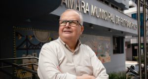 Afrânio Boppré olhando para o horizonte em frente a Câmara Municipal de Florianópolis. Ele tem 61 anos, cabelos brancos, usa óculos e veste uma camisa branca.