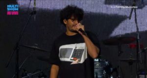 Criolo usa camiseta de urna eletrônica e plateia xinga Bolsonaro em show do Emicida no Lollapalooza. Foto do rapper utilizando a camisa e falando ao microfone.