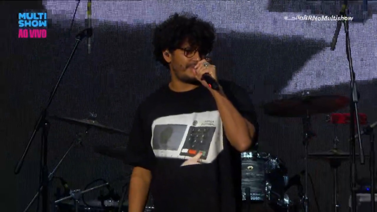 Criolo usa camiseta de urna eletrônica e plateia xinga Bolsonaro em show do Emicida no Lollapalooza. Foto do rapper utilizando a camisa e falando ao microfone.