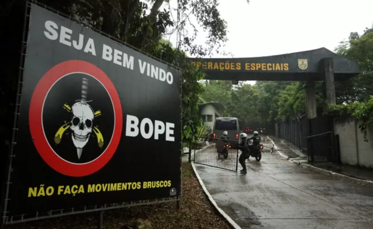 Foto da entrada do BOPE. Há uma placa dando boas vindas, onde também está escrito: "Não faça movimentos bruscos".