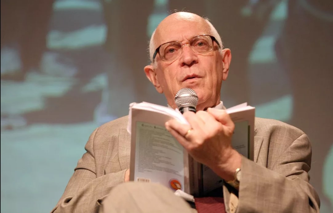 O jurista Dalmo Dallari sentado, falando ao microfone, com um livro na mão, em um evento em 2008.