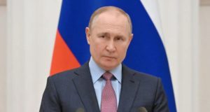 Vladmir Putin com expressão séria posando para a foto em frente a bandeira da Rússia.