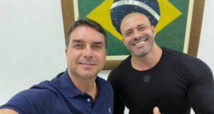 Flávio Bolsonaro e Daniel Silveira posando para a foto com uma bandeira do Brasil atrás deles.