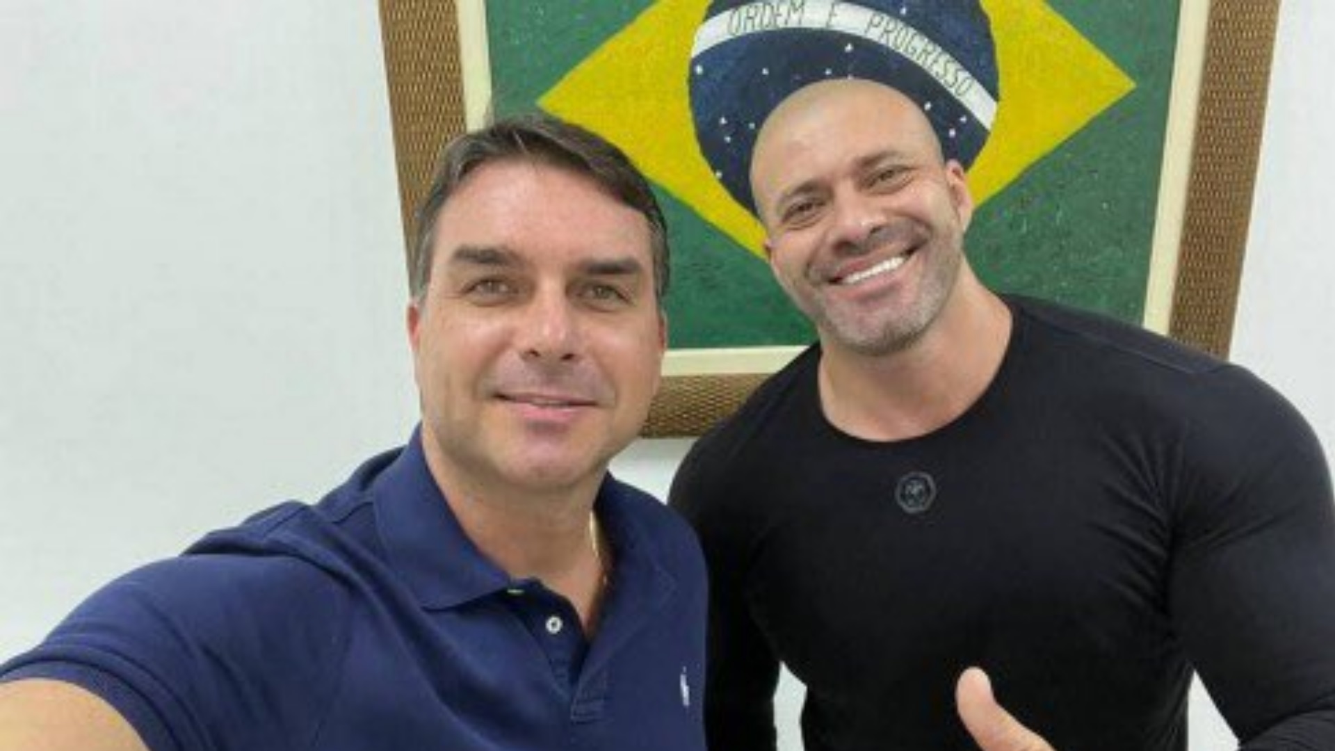 Flávio Bolsonaro e Daniel Silveira posando para a foto com uma bandeira do Brasil atrás deles.