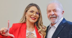 Deolane aparece com roupa vermelha, cabelos loiros longos, óculos e sorri. Ao lado, o ex-presidente Lula aparece usando terno preto.