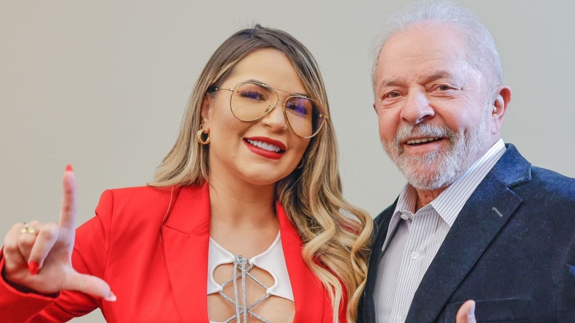 Deolane aparece com roupa vermelha, cabelos loiros longos, óculos e sorri. Ao lado, o ex-presidente Lula aparece usando terno preto.