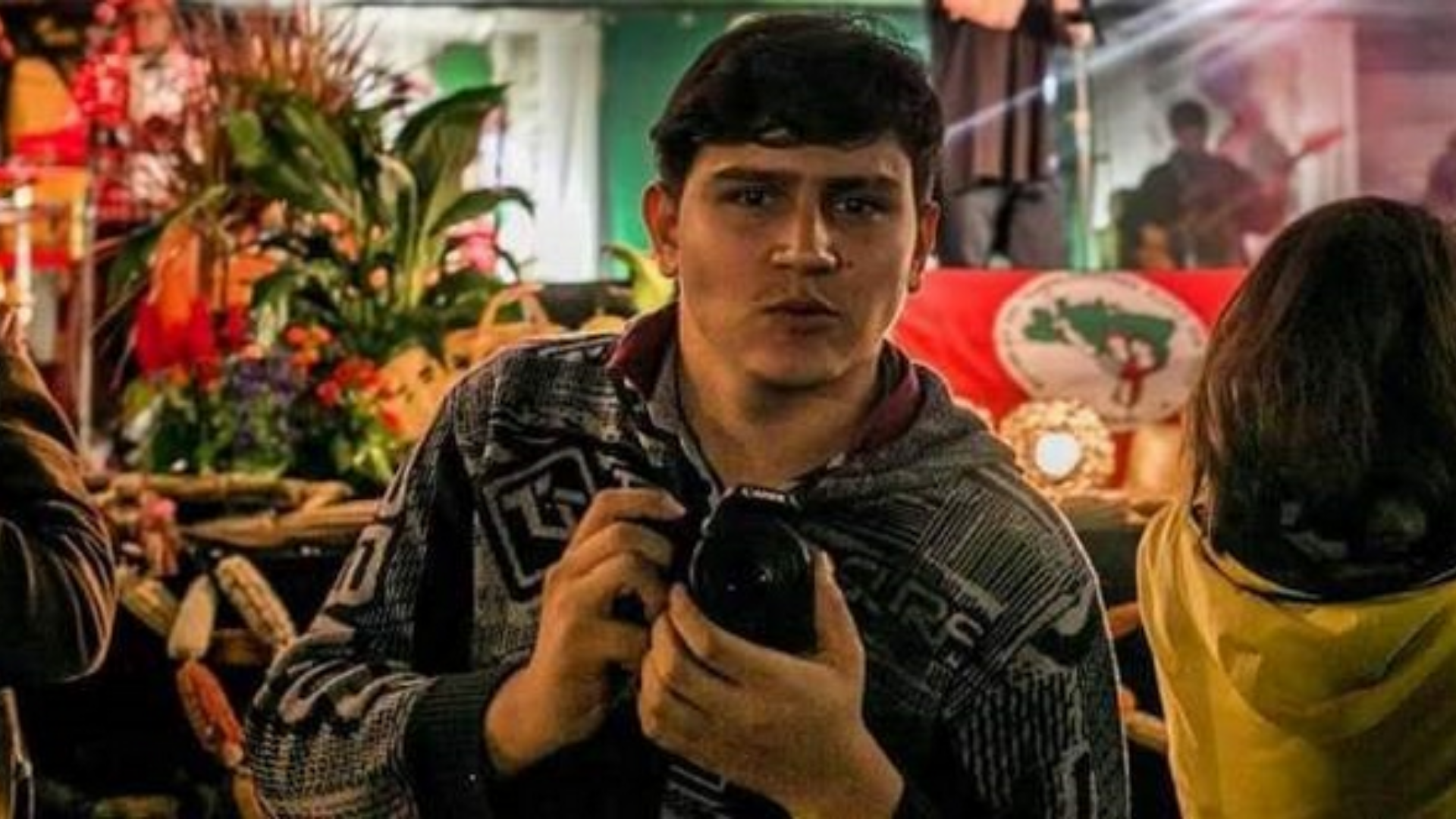 Jornalista promete entrar na Justiça contra Luciano Hang após sofrer ataques na web. Foto do fotojornalista com uma câmera, cabelo preto e uma bandeira do MST ao fundo.