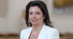 Foto de Margarita Simonyan usando um vestido branco, sorrindo, cabelos pretos lisos e penteados. ela usa brincos e tem pele branca.