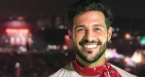 Nova atualização do estado clínico do ex-BBB Rodrigo Mussi é divulgada. Foto de Rodrigo Mussi sorrindo com um cachecol vermelho.