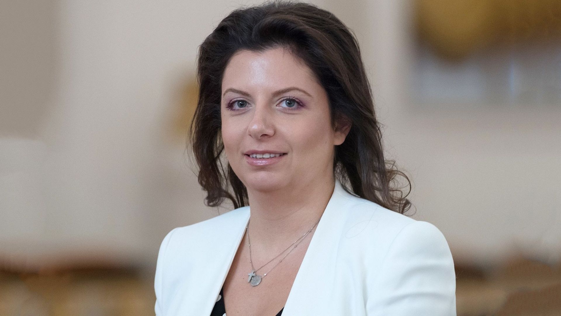 Foto de Margarita Simonyan usando um vestido branco, sorrindo, cabelos pretos lisos e penteados. ela usa brincos e tem pele branca.