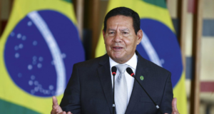 Foto de Hamilton Mourão em um púlpito falando ao microfone. Ao lado, há uma bandeira do Brasil.