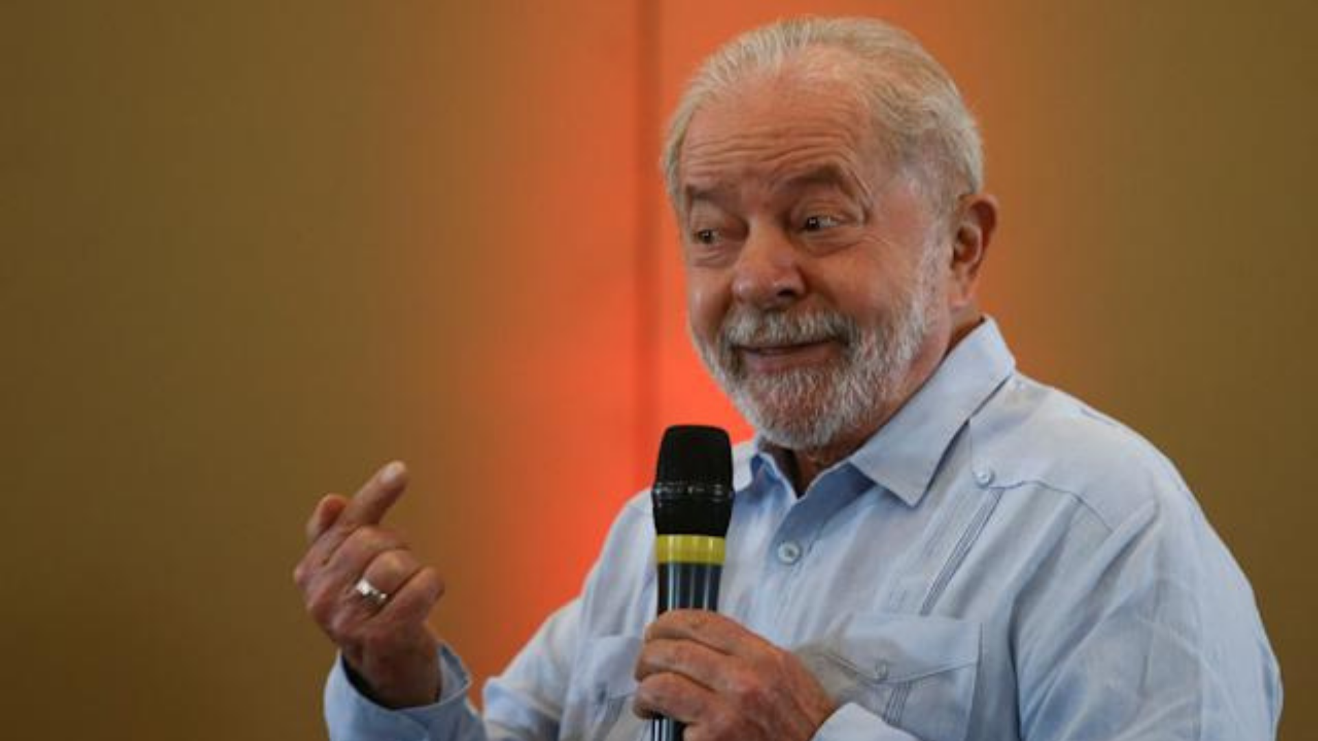Foto do ex-presidente Lula falando ao microfone com um sorriso no rosto.