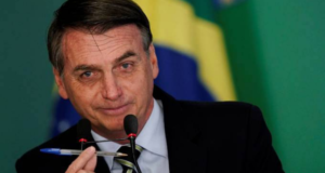 Bolsonaro aparece sorrindo enquanto fala ao microfone e segura uma caneta com a mão.