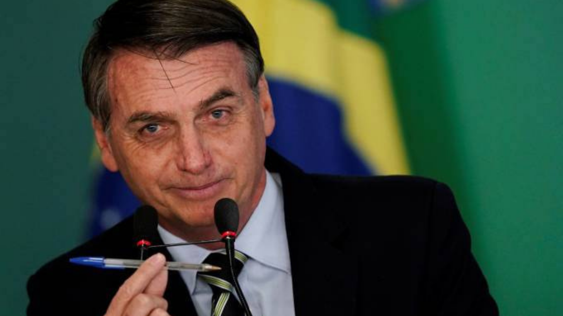 Bolsonaro aparece sorrindo enquanto fala ao microfone e segura uma caneta com a mão. PGR decide que não irá investigar o presidente.