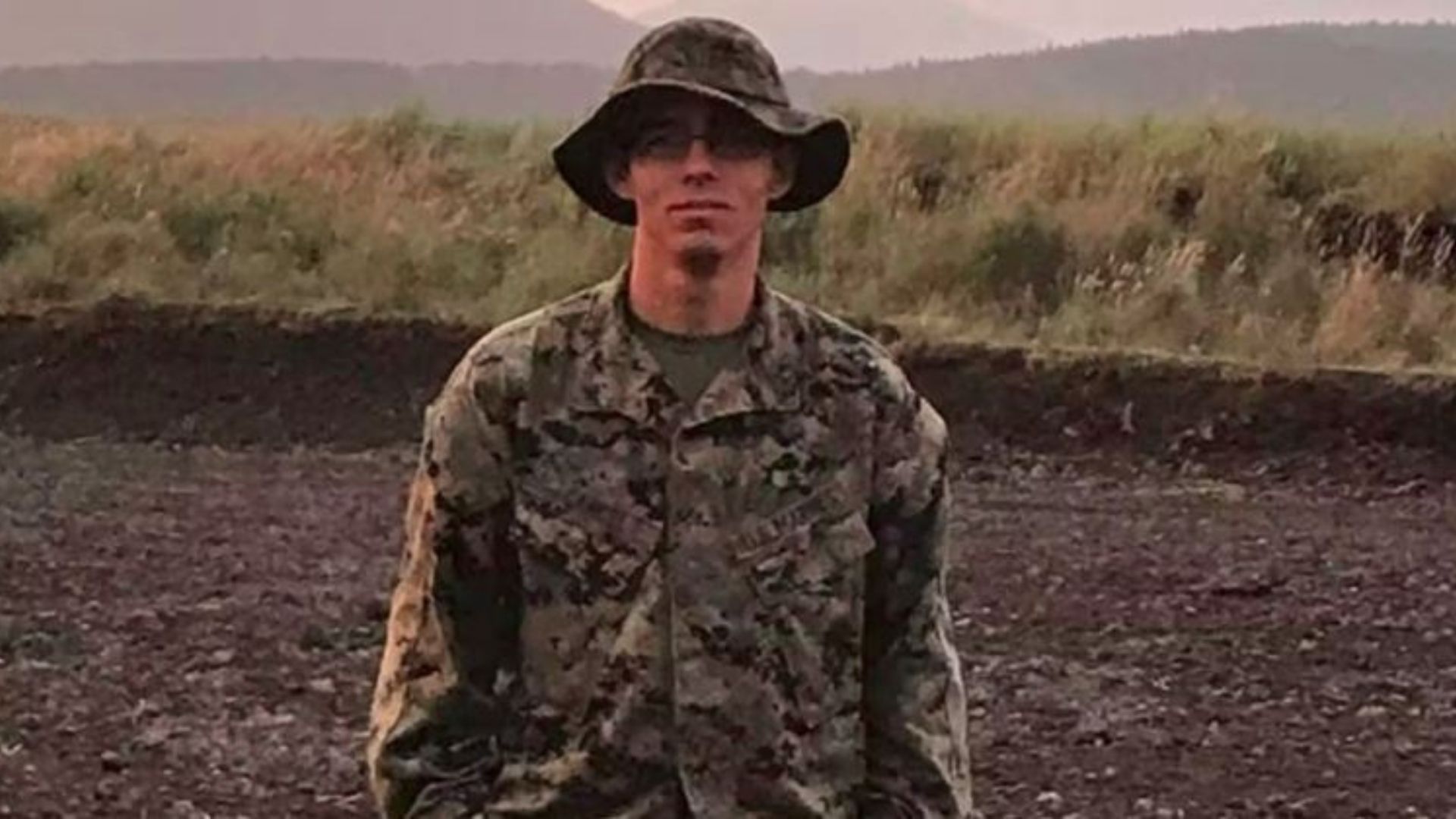foto de Cancel com uma roupa tradicional do exército, barba rala no queixo e paisagem verde e montanhosa ao fundo.