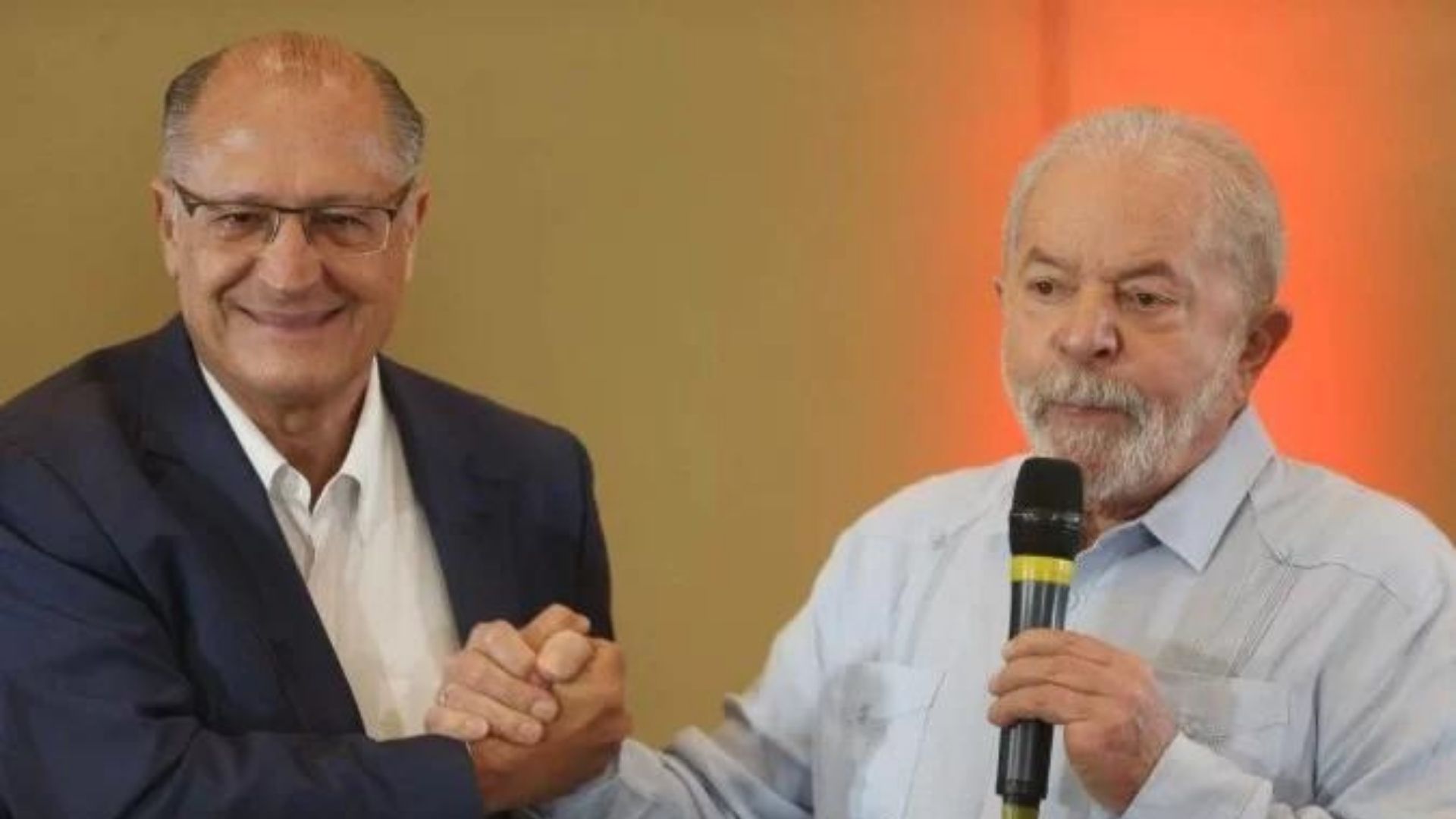 Lula e alckmin aparecem em aperto de mãos em um evento.
