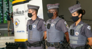Tarcisio Freitas diz que o equipamento nada resolve. Policiais militares utilizando câmeras no fardamento. Eles estão em pé, utilizando máscaras e com expressão séria.
