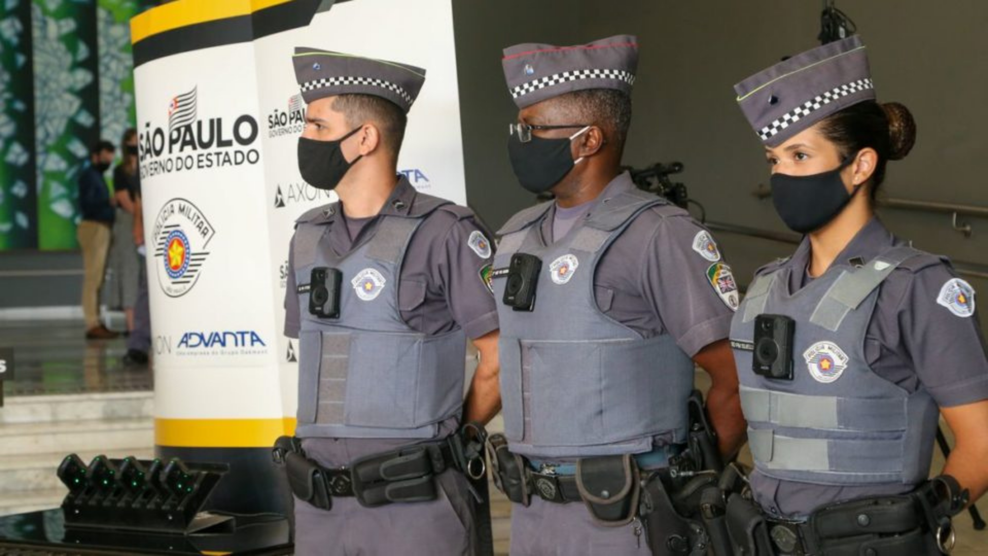 Tarcisio Freitas diz que o equipamento nada resolve. Policiais militares utilizando câmeras no fardamento. Eles estão em pé, utilizando máscaras e com expressão séria.