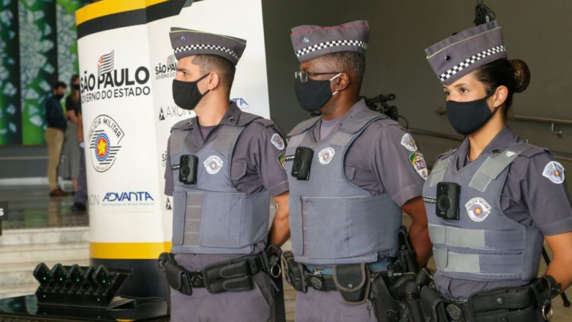 Tarcisio Freitas diz que o equipamento nada resolve. Policiais militares utilizando câmeras no fardamento. Eles estão em pé, utilizando máscaras e com expressão séria. 