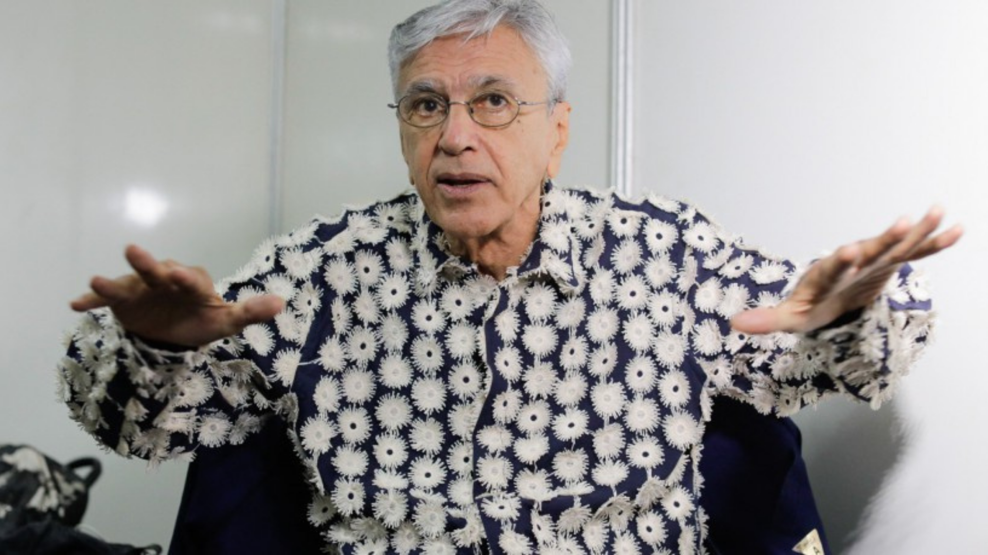 Caetano Veloso irá processar Bolsonaro. O cantor aparece com uma camisa branca e bolinhas pretas, com as duas mãos no ar.