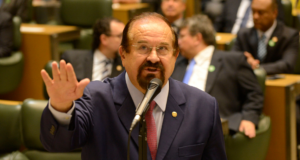 Aldo Demarchi faz juamento com a mão direita, fala ao microfone e usa terno azul marinho.