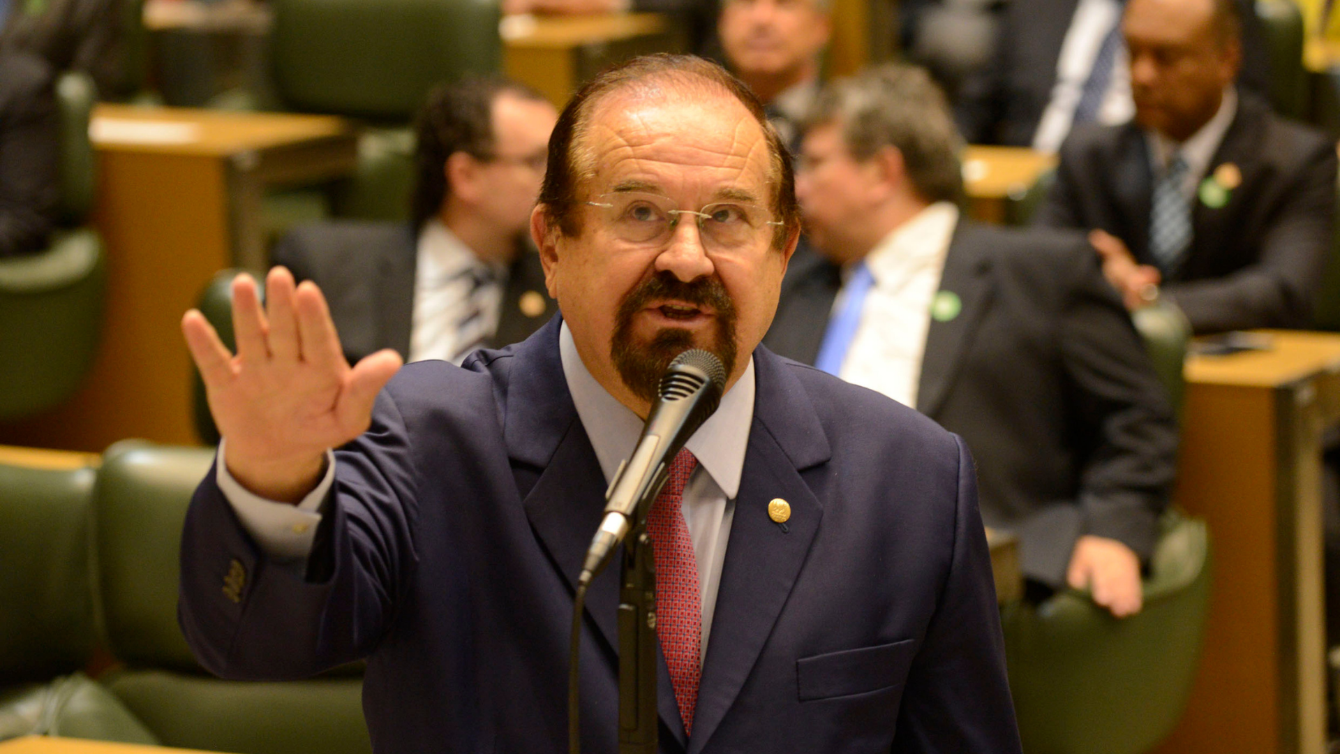 Aldo Demarchi faz juamento com a mão direita, fala ao microfone e usa terno azul marinho.