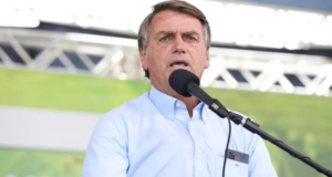 Bolsonaro aparece discursando em um microfone em um púlpito com um fundo verde.