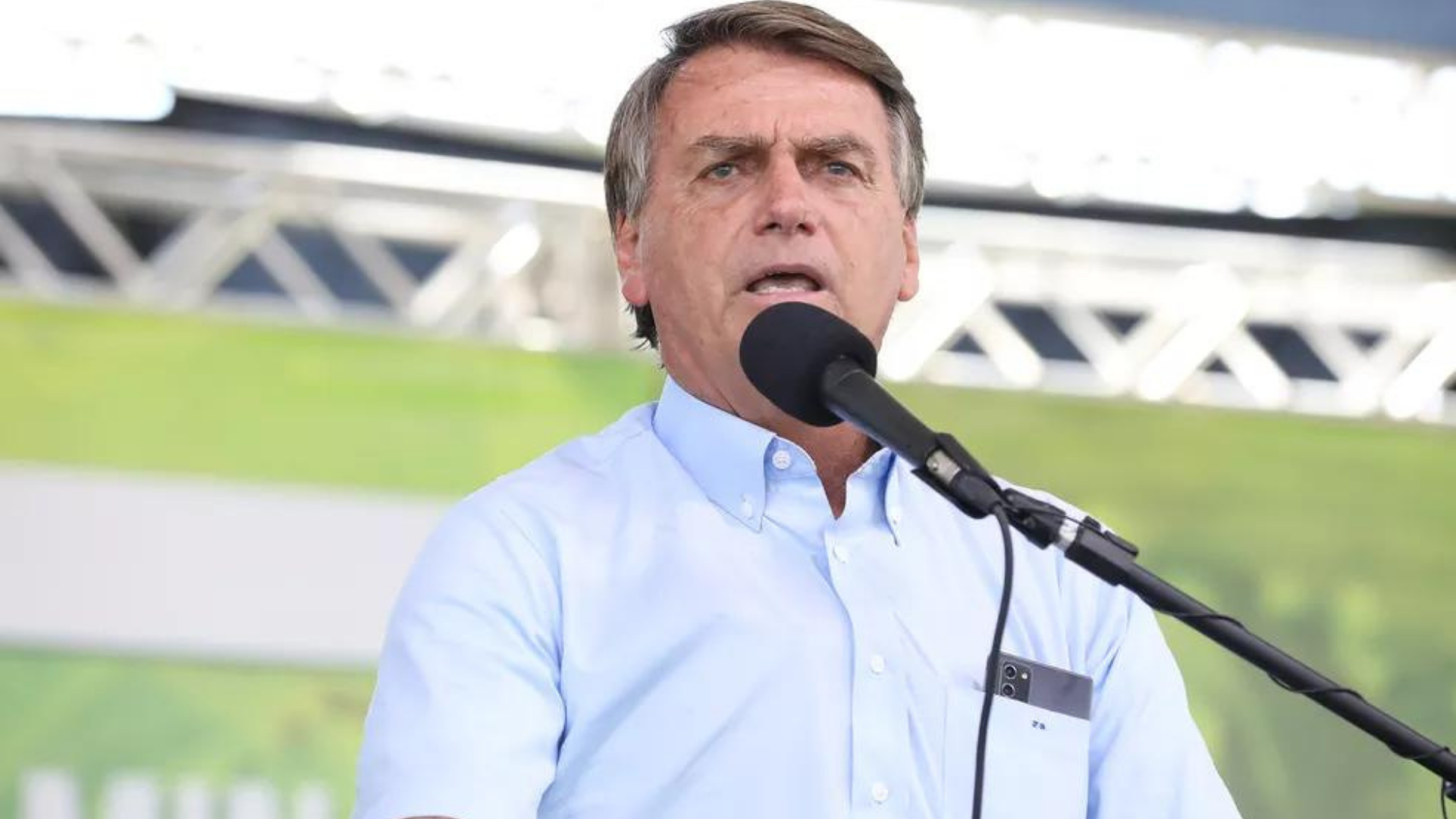 Bolsonaro aparece discursando em um microfone em um púlpito com um fundo verde.