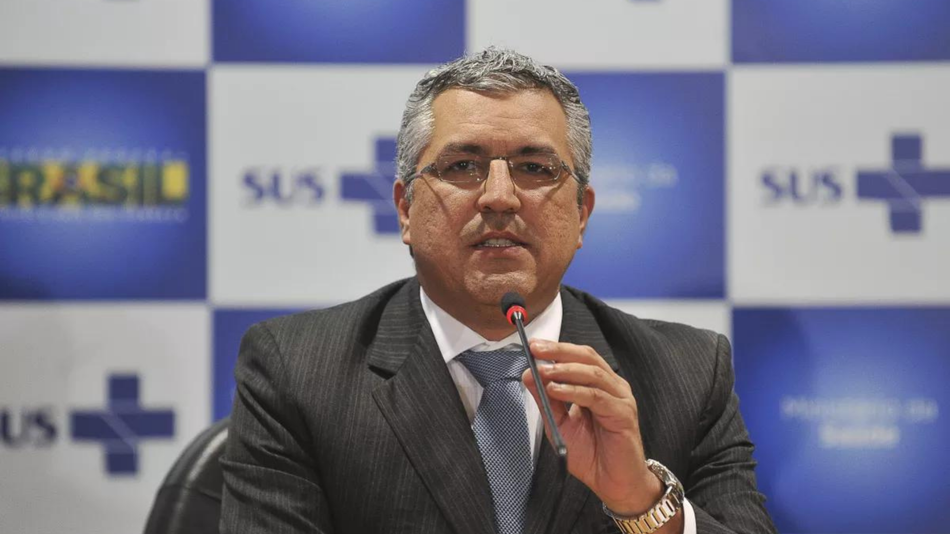 Foto de Alexandre Padilha usando óculos, cabelo grisalho, terno preto e gravata. 