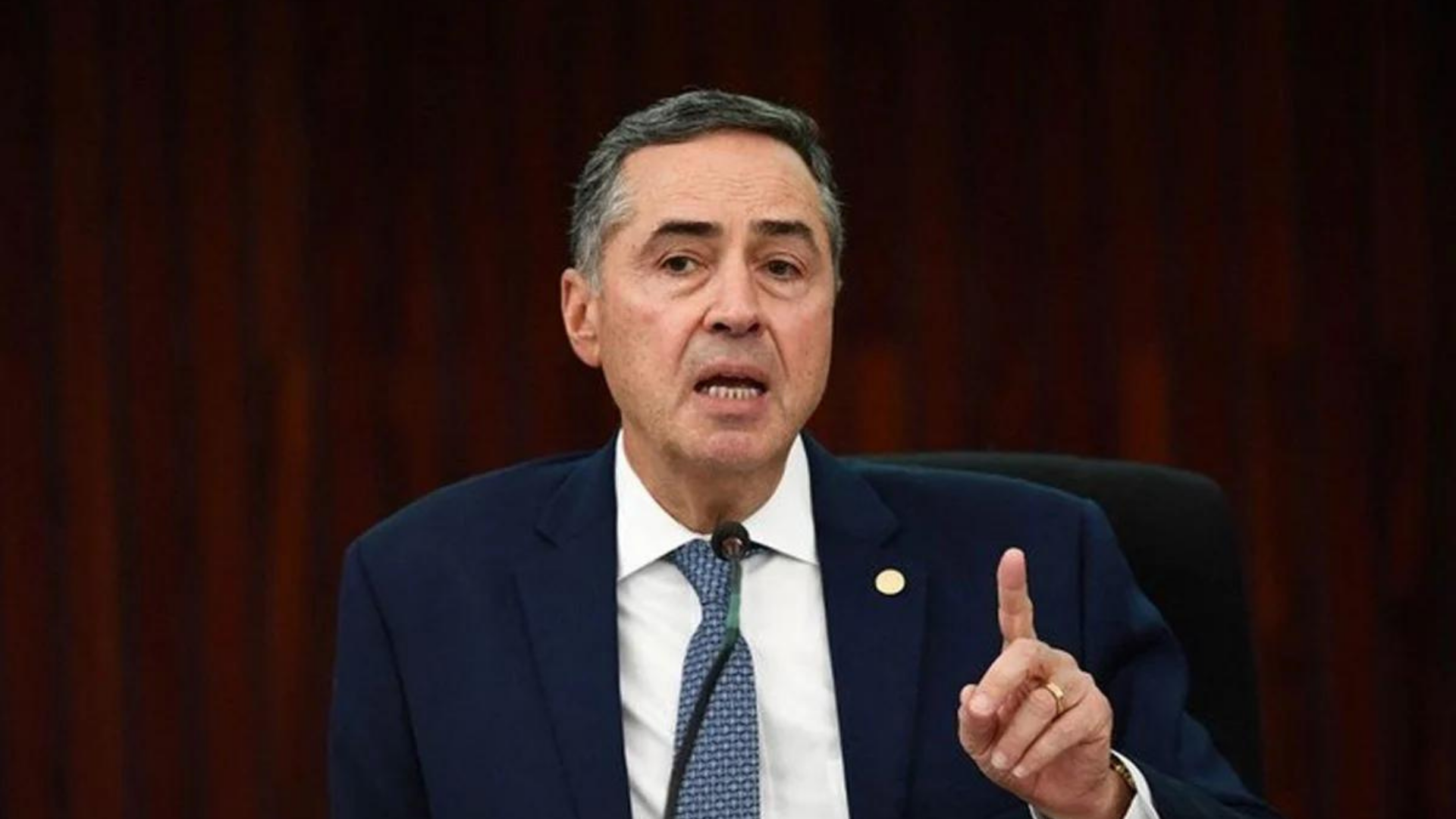 Foto de Luís Roberto Barroso com olhar sério e apontando o dedo indicador esquerdo para cima.