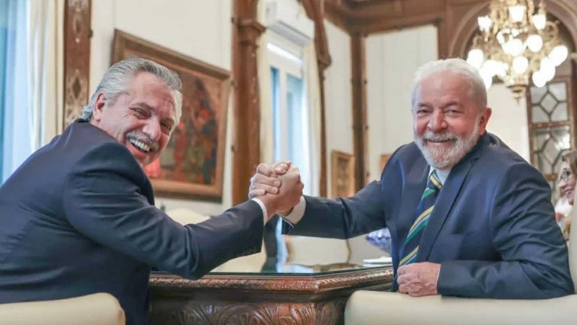 Foto do ex-presidente Lula ao lado de Alberto Fernández. Eles estão sentados, sorriem em um aperto de mãos.