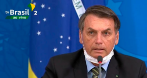 Bolsonaro fala ao micrfoone com máscara no queixo.