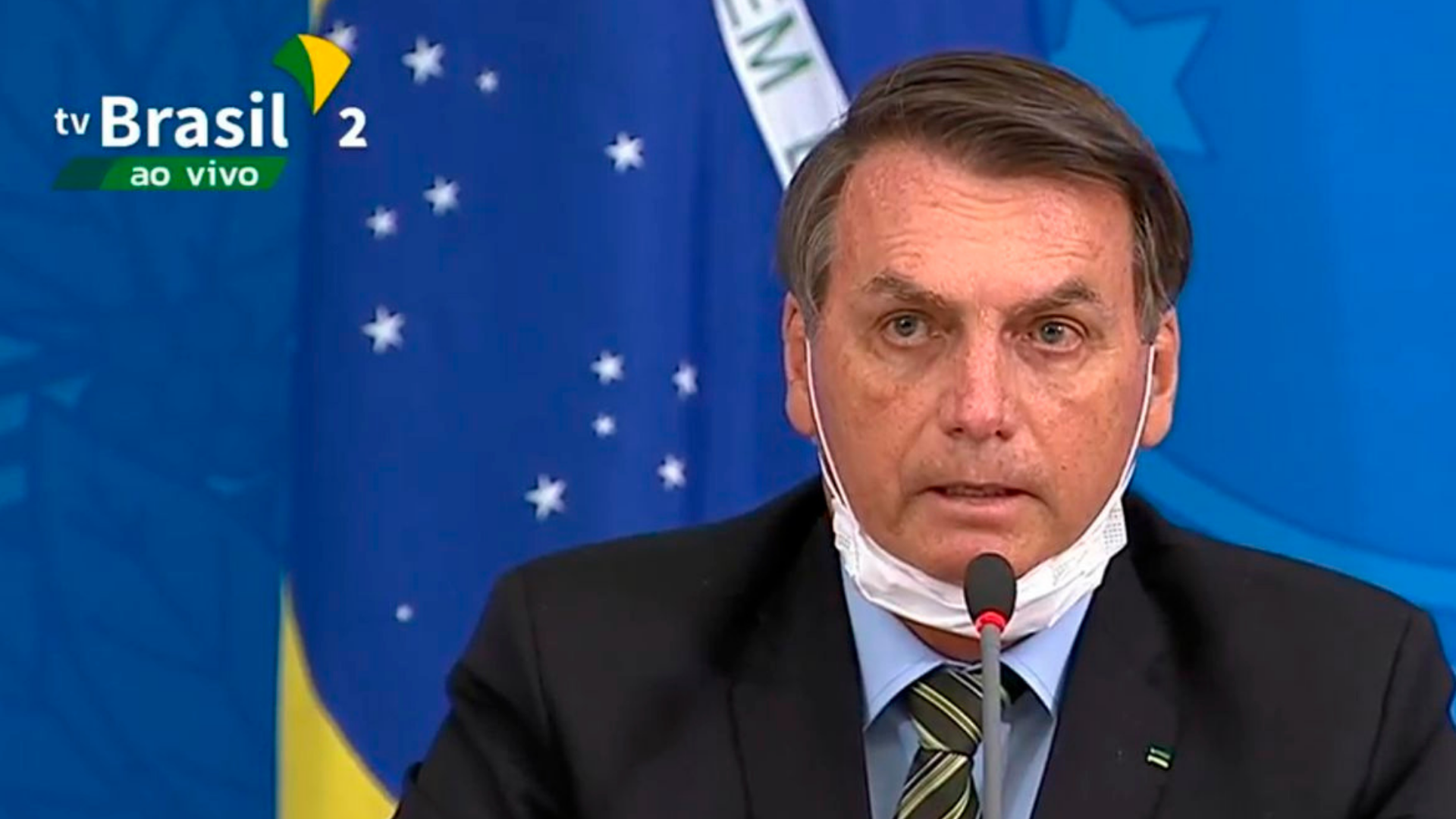 Bolsonaro fala ao micrfoone com máscara no queixo.