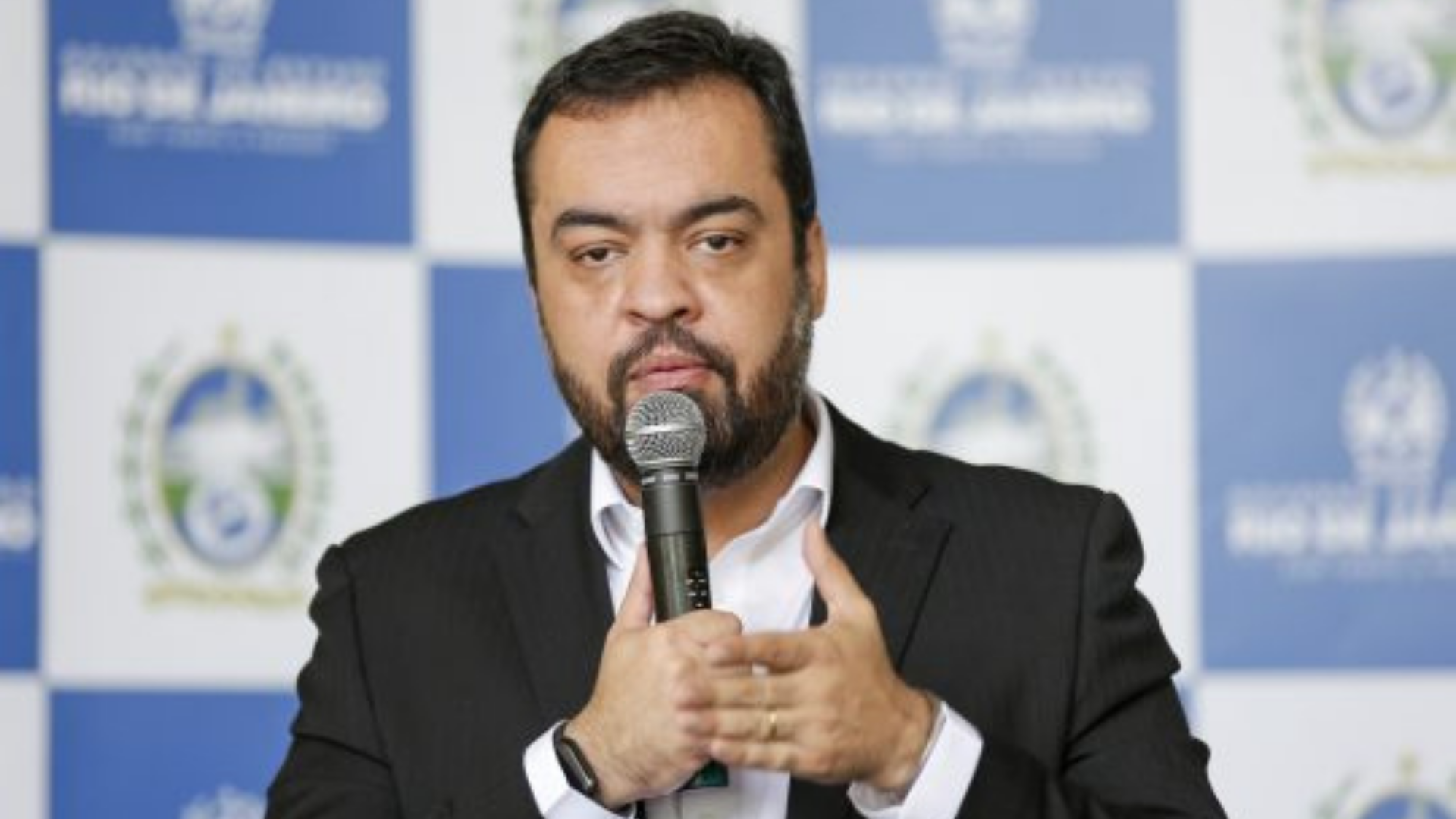 Helicóptero com governador do Rio faz pouso de emergência. Foto d eCláudio Castro falando ao microfone e com terno preto.