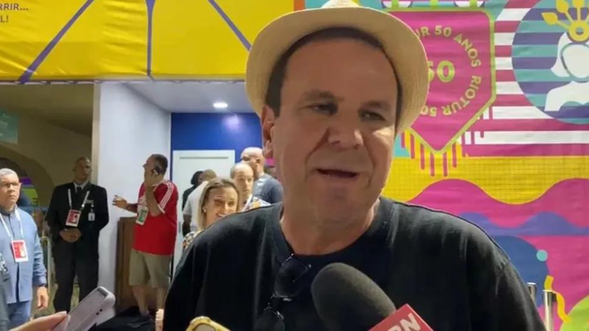 Foto de Eduardo Paes no carnaval do Rio falando ao microfone. ele usa um chapéu enquanto concede entrevista.