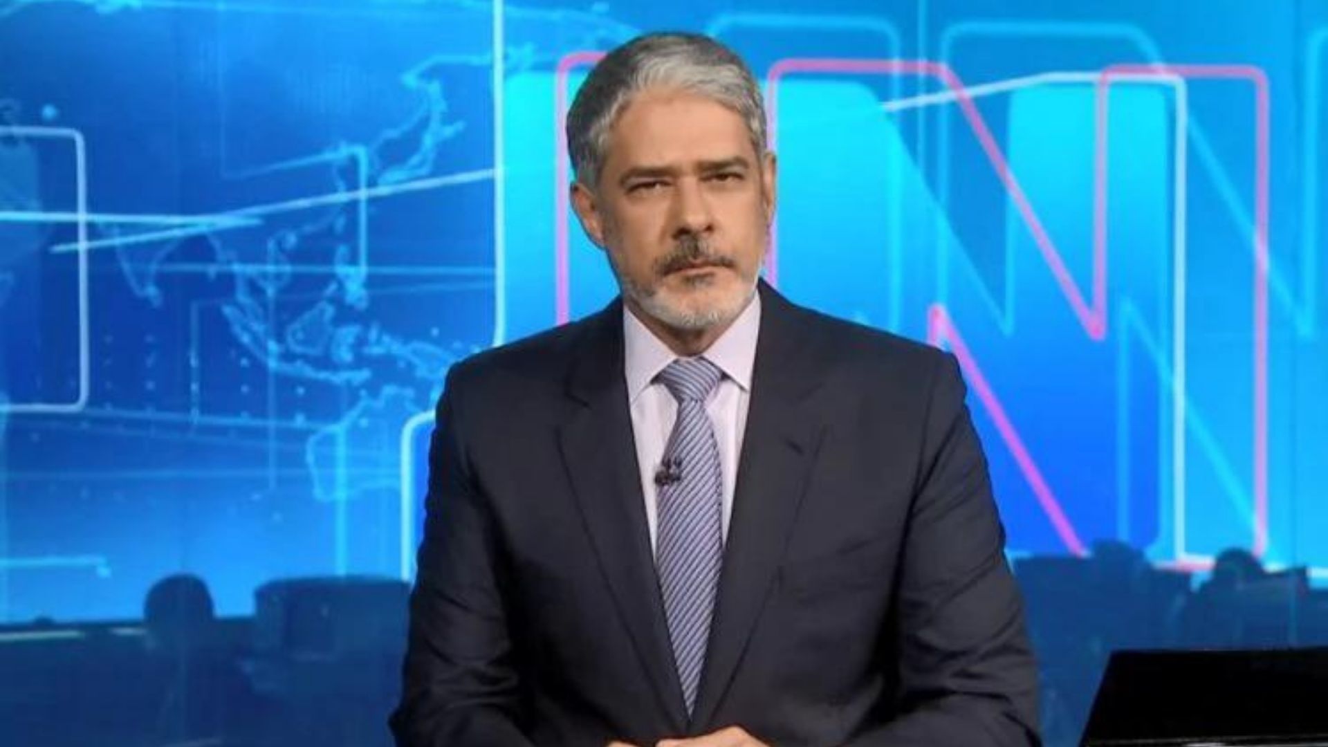 William Bonner usa terno preto, gravata, barba e cabelos grisalhos. Ele está no estúdio do Jornal Nacional.