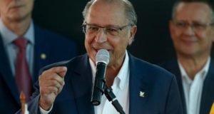 Alckmin fala ao microfone em um evento. Ele usa terno preto, óculos e é calvo.