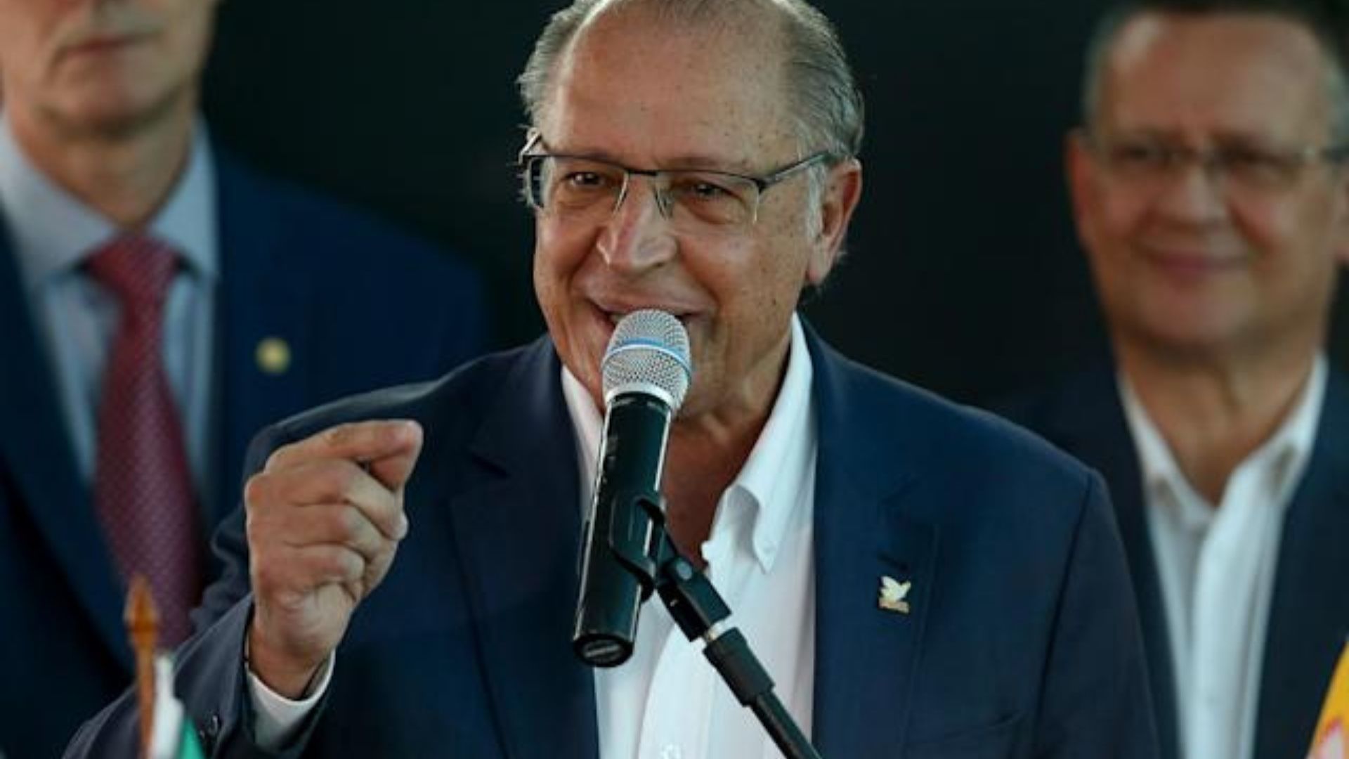 Alckmin fala ao microfone em um evento. Ele usa terno preto, óculos e é calvo.