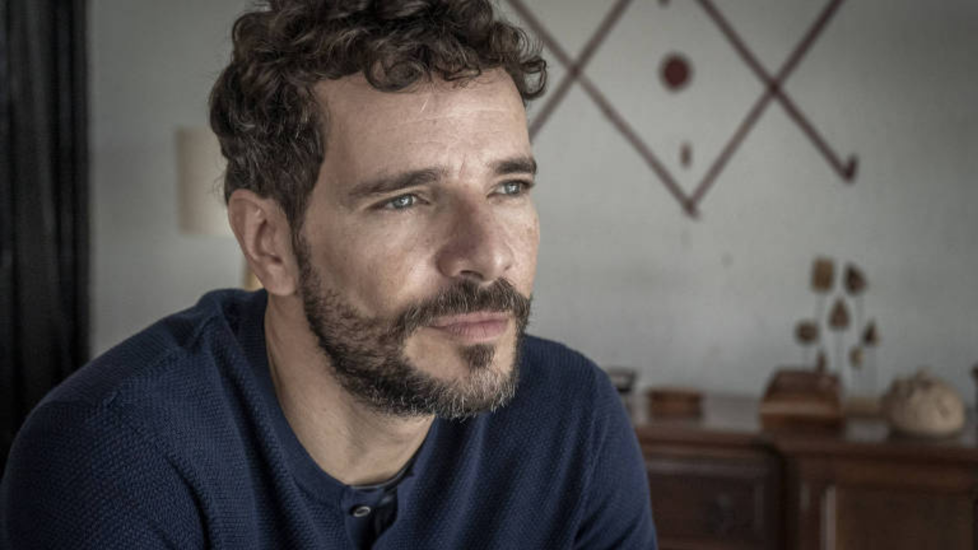 Daniel de Oliveira deixará a Globo. Ele tem cabelo e barba grisalhos. Ele tem expressão séria e olha para um ponto distante.