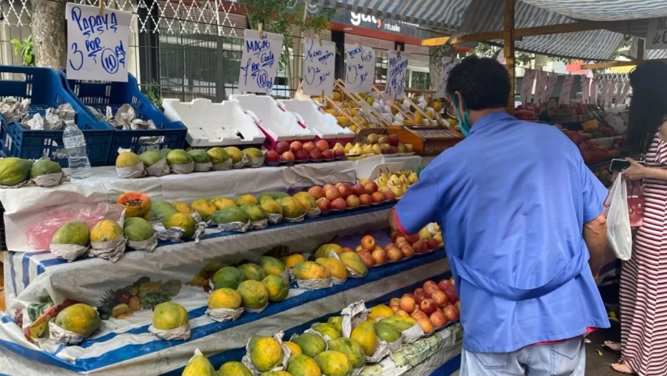 Imagem ilustrativa de uma feira. Uma pessoa aparece comprando frutas.
