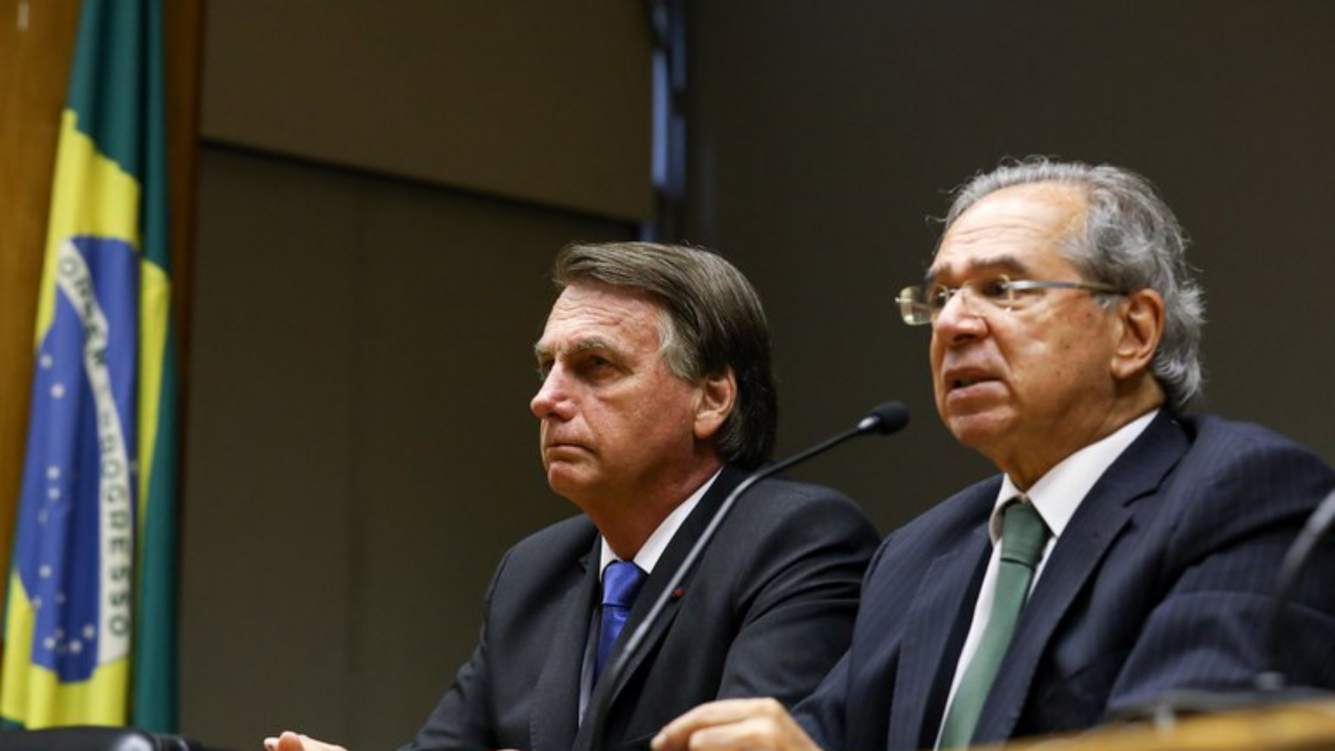 Foto de Bolsonaro ao lado de Paulo Guedes, ambos usam terno e estão sentados.