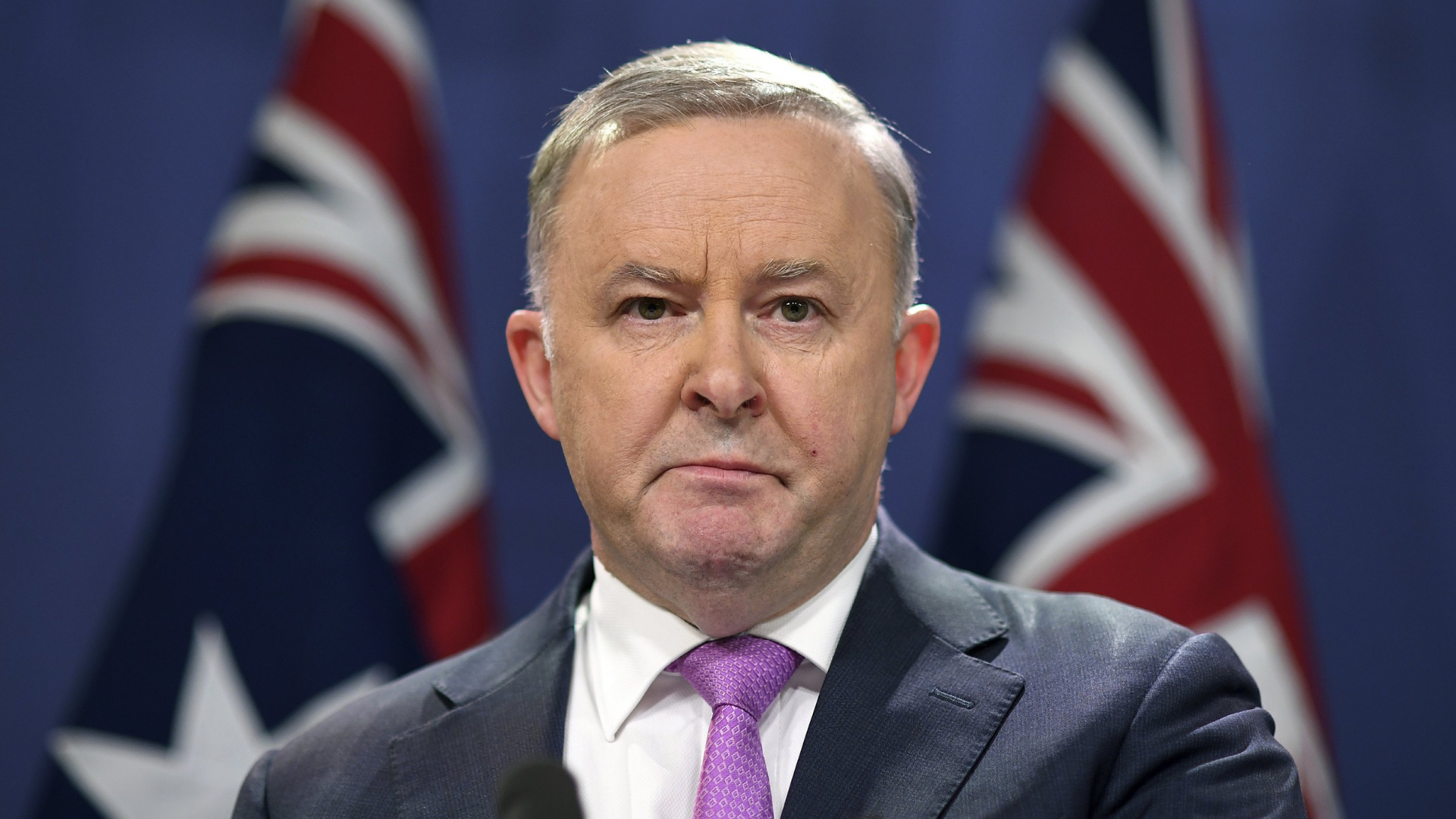 foto de anthony Albanese, com terno preto, gravata lilás, cabelo branco e bandeiras da Austrália desfocadas ao fundo.