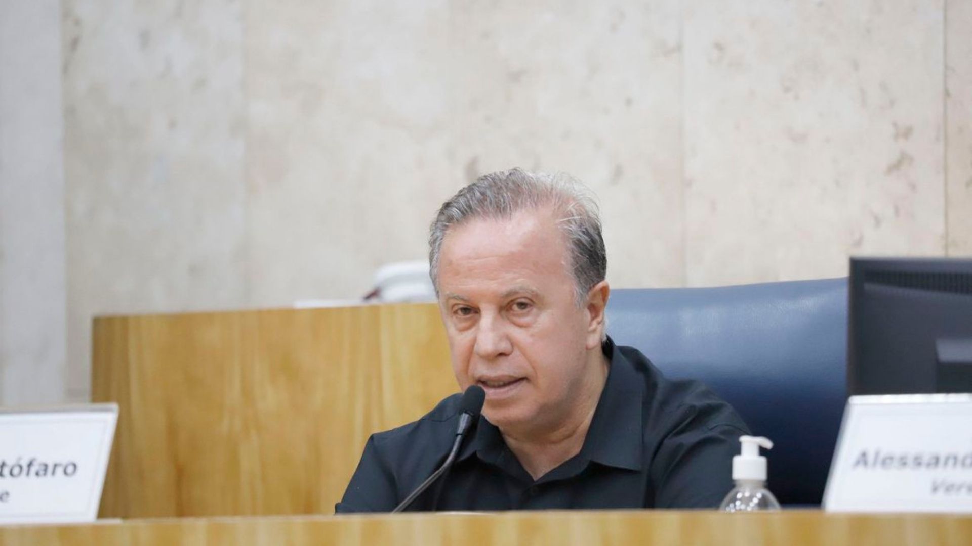 Foto de Camilo cristófaro sentado em um plenário. Ele usa camisa preta, fala ao microfone, tem cabelo lambido e expressão séria.