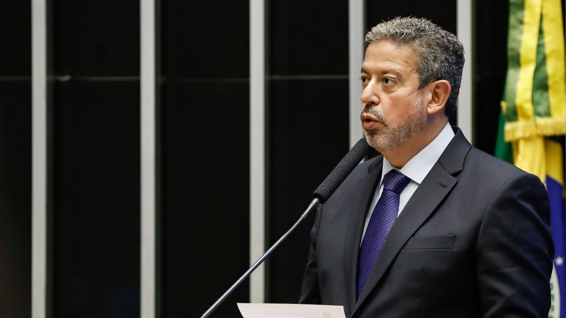 Foto de Arthur Lira falando ao microfone na Câmara dos Deputados. Ele usa terno preto, gravata azul, pele branca, barba rala e cabelo grisalho.