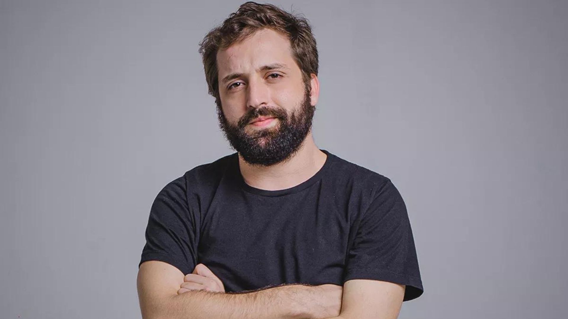 Gregório Duvivier usando camisa preta, barba, pele branca e sorriso. Parede branca ao fundo. 