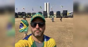 Márcio Furtado usa boné verde, ´óculos de sol, camisa cinza, bandeira do Brasil, barba rala e prédio do Congresso ao fundo.