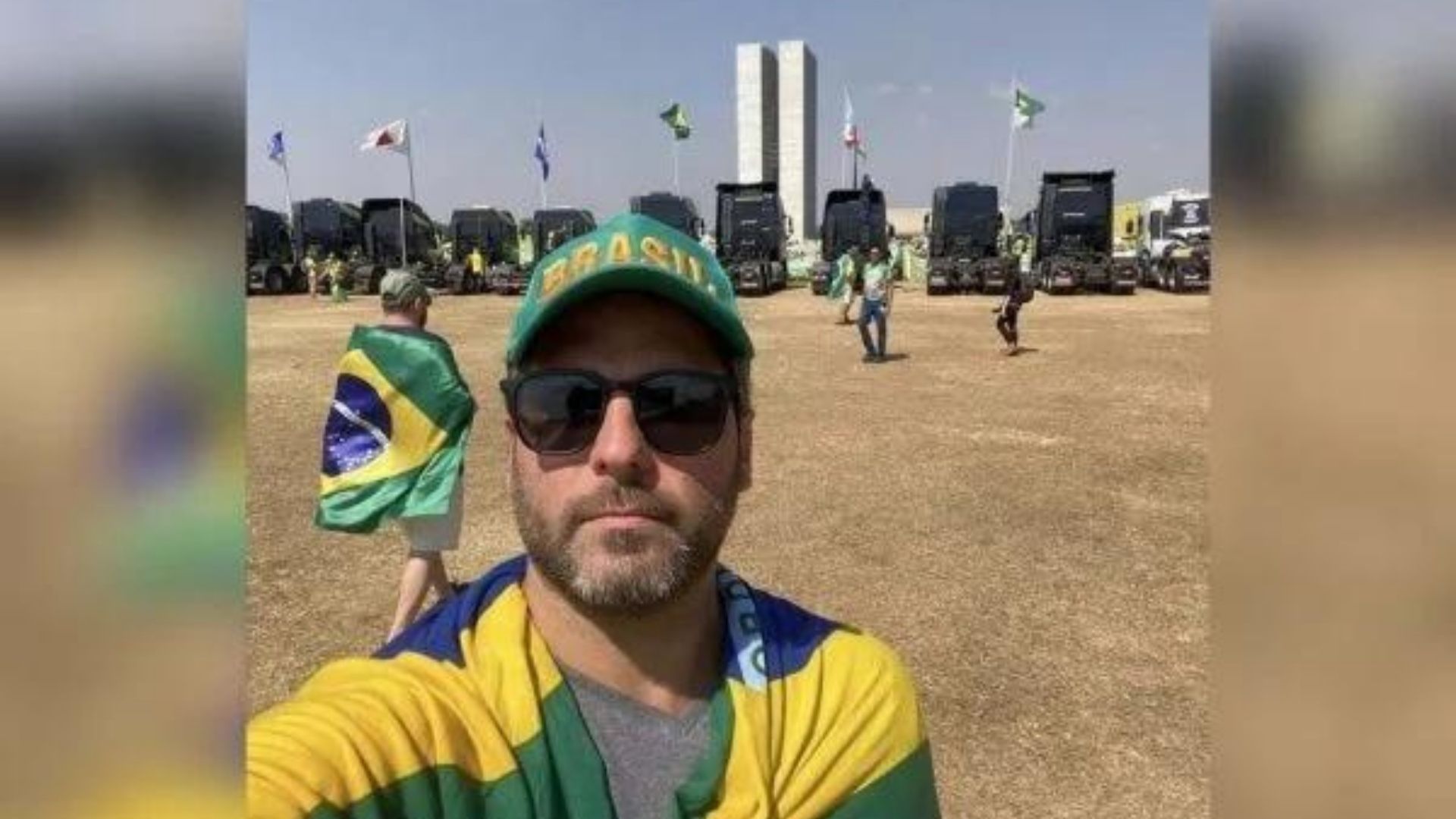 Márcio Furtado usa boné verde, ´óculos de sol, camisa cinza, bandeira do Brasil, barba rala e prédio do Congresso ao fundo.